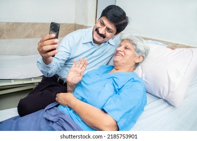 Indian Old Senior Patient In Hospital Bed Using Smart Phone Doing Video Call To Descendant Relatives With Son Sitting By Side - Elderly Medical And Healthcare Concept