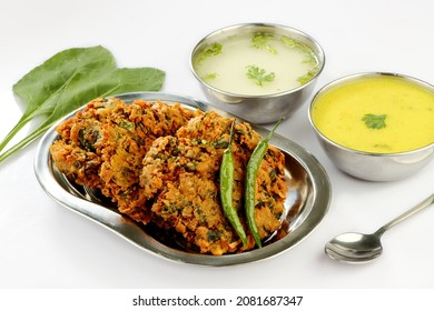 Indian Popular Snack Called Palak Vada Or Spinach  Fritter In Steel Plate And Yellow Souse Called Kadhi In Bowl White Souse Called Mattha In Bowl And Green Chilli On White Background 