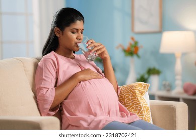 Indian pregnant woman drinking water while sitting on sofa at home - concept of hydration, healthy pregnancy lifestyle and wellness - Powered by Shutterstock