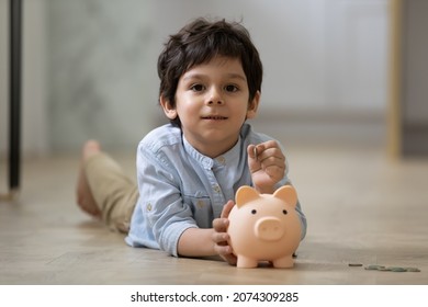 Indian preschool boy putting coin into pink piggy bank, playing game, resting on warm floor at home. Little kid saving money, cash, learning economy, planning future investment. Financial education