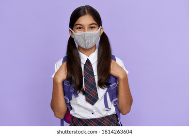 Indian preteen girl, latin kid schoolgirl student wears uniform and face mask for coronavirus protection safety holding backpack stands isolated on lilac violet background looking at camera, portrait.