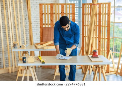 Indian professional bearded male engineer architect foreman labor worker carpenter wears safety helmet and gloves using wood polishing machine grinding sanding wooden board surface on workbench.