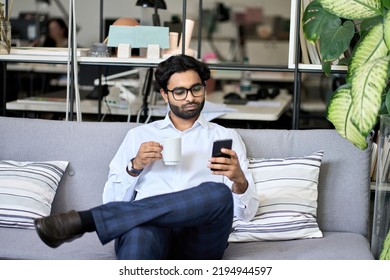 Indian Professional Business Man Executive Holding Smartphone Using Mobile Phone Reading News, Checking Financial Market Or Planner Applications Sitting On Sofa Drinking Coffee Working In Office.