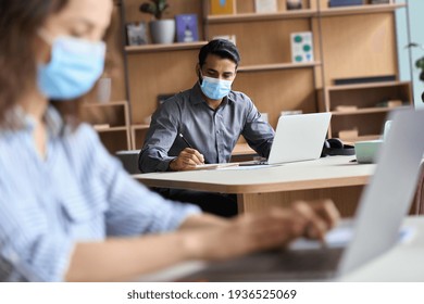 Indian professional business man or student wearing medical face mask working, studying on laptop among diverse people sitting at table in modern office coworking space keeping safe social distance.
