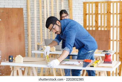 Indian Professional Male Engineer Architect Foreman Labor Worker Wears Safety Goggles Using Measuring Tape Measure Wood Plank On Working Table While Female Colleague Typing Laptop Computer Behind.