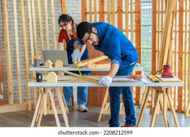 Indian Professional Male Engineer Architect Foreman Labor Worker Wears Safety Goggles Using Measuring Tape Measure Wood Plank On Working Table While Female Colleague Typing Laptop Computer Behind.