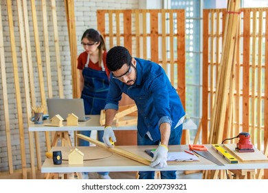 Indian Professional Male Engineer Architect Foreman Labor Worker Wears Safety Goggles Using Measuring Tape Measure Wood Plank On Working Table While Female Colleague Typing Laptop Computer Behind.