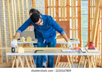 Indian Professional Male Engineer Architect Foreman Labor Worker Wears Safety Goggles Using Measuring Tape Measure Wood Plank On Working Table While Female Colleague Typing Laptop Computer Behind.