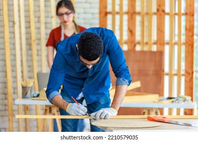 Indian Professional Male Engineer Architect Foreman Labor Worker Wears Safety Goggles Using Measuring Tape Measure Wood Plank On Working Table While Female Colleague Typing Laptop Computer Behind.