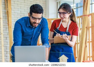 Indian Professional Male Engineer Foreman Labor Worker Using Laptop Computer Meeting Discussing With Cheerful Female Colleague Wear Safety Goggles Writing Note On Paper Clipboard In Construction Site.