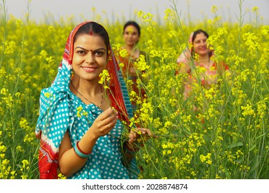 Indian Rural Villager Woman Farmer Standing Happily In A Mustard Field In A Green Farm Yellow Flowers Enjoying The Thriving Agricultural Crops In Prosperity. Women Standing In The Background