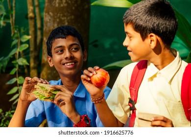 Indian School Boy Eating Sandwich And His Friend With Backpack Sits On Table And Eating An Apple In Park