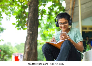 Indian School Boy Holding Phone Distance Learning Class Using Mobile Application, Watching Online Lesson, Video Calling In App Making Notes Studying At Home