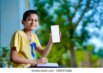 Indian School Boy Holding Phone Distance Learning Class Using Mobile Application, Watching Online Lesson, Video Calling In App Making Notes Studying At Home