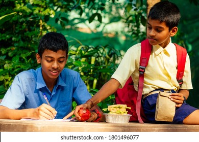 Indian School Boy Writing On Notepad Doing Homework , Looking Concentration . His Friend With Backpack Sits On Table And Eating An Apple In Park