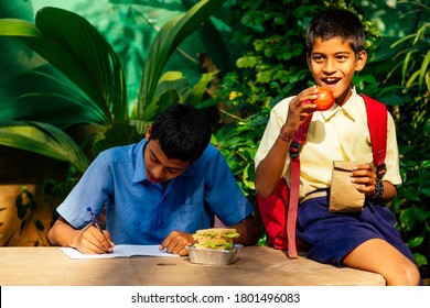 Indian School Boy Writing On Notepad Doing Homework , Looking Concentration . His Friend With Backpack Sits On Table And Eating An Apple In Park