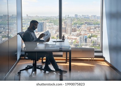 Indian serious investment banker financial analyst sitting at desk with papers working with big data, stock exchange trading operations using laptop near panoramic window in modern corporate office.