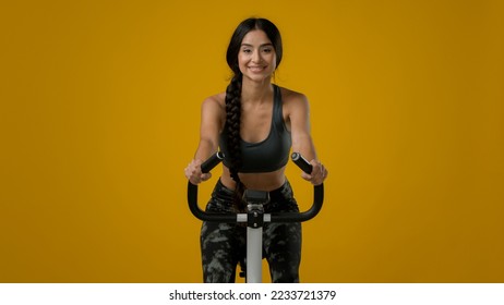 Indian slim happy smiling sporty woman female lady athletic girl cyclist biking flexing cycling riding on exercise bike sport equipment fitness workout cardio ride in yellow studio thumb up gesture - Powered by Shutterstock