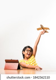 Indian Small Girl Student Making Or Measuring Wings Of Paper Plane Or Aeroplane, Doing School Project Or Learning Science With Online Tutorial