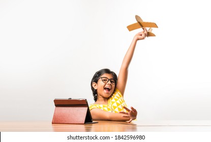 Indian Small Girl Student Making Or Measuring Wings Of Paper Plane Or Aeroplane, Doing School Project Or Learning Science With Online Tutorial