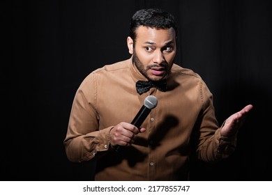 Indian Stand Up Comedian Gesturing While Telling Jokes Into Microphone On Dark Stage Isolated On Black