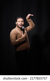 Indian Stand Up Comedian Gesturing While Telling Joke Into Microphone On Dark Stage On Black