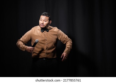 Indian Stand Up Comedian Grimacing While Telling Joke Into Microphone On Dark Stage On Black