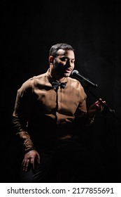 Indian Stand Up Comedian Telling Jokes Into Microphone On Dark Stage Isolated On Black