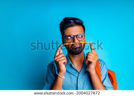 indian student man feeling boring wear eye glasses holding a pencil over lip like a mustache in blue studio