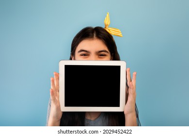 Indian Teen Girl Hiding Behind Tablet Computer With Empty Screen On Blue Studio Background, Mockup For Website Or App. Cool Adolescent Showing Touch Pad With Space For Ad