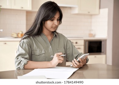 Indian teen girl holding mobile phone using remote online education app at home. Female student using smartphone online learning in application program writing notes sit at table at home office.