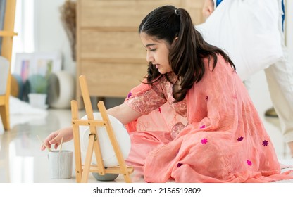 Indian Teen Girl Wearing Traditional Clothes, Playing Alone, Using Brush Painting Art On Plate In Leisure Time With Happiness, Sitting On Floor In Living Room At Home. Lifestyle, Education Concept