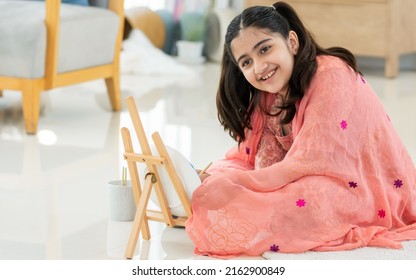 Indian Teen Girl Wearing Traditional Clothes, Playing Alone, Using Brush Painting Art On Plate In Leisure Time With Happiness, Sitting On Floor In Living Room At Home. Lifestyle, Education Concept