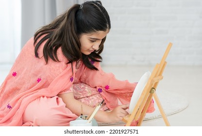 Indian Teen Girl Wearing Traditional Clothes, Playing Alone, Using Brush Painting Art On Plate In Leisure Time With Happiness, Sitting On Floor In Living Room At Home. Lifestyle, Education Concept