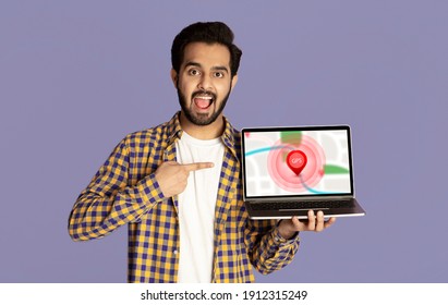 Indian Tourist Man Showing Laptop Computer With Online Maps For Navigation Posing Standing Over Purple Studio Background, Smiling To Camera. Online GPS Navigator Service.