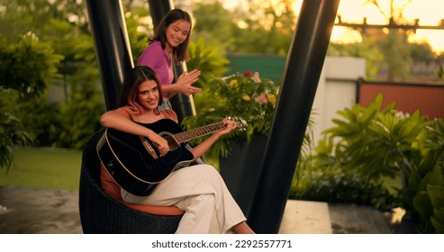 Indian two girl sitting on chair singing playing acoustic guitar having fun with music. Beautiful smiling young ladies couple relax enjoy practicing musical instrument spend evening time together  - Powered by Shutterstock