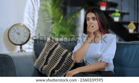 Indian Unhappy tense female sitting on sofa couch watching shocked bad news on TV at indoor house. Beautiful lonely scared sad woman feeling panic suffer stress thinking negative at home