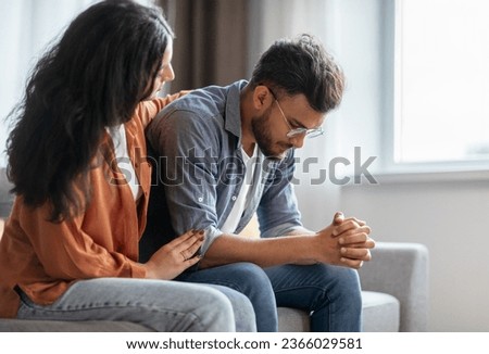 Indian woman calming upset offended guy, support and caring, sitting on sofa at home, free space. Sorry after quarrel, relationship problems, bad news reaction and crisis