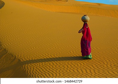 Indian Woman Carrying Heavy Jug Of Water On Her Head And Walking On A Yellow Sand Dune In The Hot Summer Desert Against Blue Sky.water Crises, Jaisalmer, Rajasthan, India