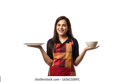 Indian Woman Chef Wearing Apron And Holding Empty Ceramic Dinner Plate And Bowl With Different  Facial Expressions, Standing Isolated Over White Background