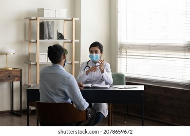 Indian Woman Doctor Physician In Protective Medical Mask Consulting African American Man Patient, Sitting At Desk In Hospital, Female Practitioner Discussing Checkup Or Symptoms, Coronavirus Concept