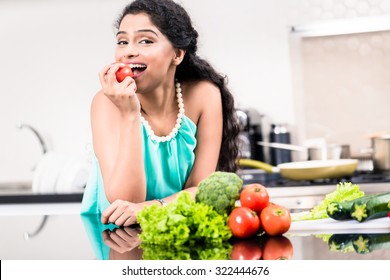 Indian Woman Eating Healthy Apple In Her Kitchen, Salad And Vegetables On The Counter