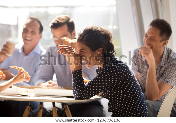 Indian woman laughing at funny joke eating pizza\
with diverse coworkers in office, friendly work team enjoying\
positive emotions and lunch together, happy colleagues staff group\
having fun at break