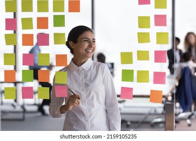 Indian woman office employee create visual plan for colleagues, prepare for briefing, develop strategy, make notes use colorful sticky notes attached on glass wall smile looks aside. Workflow concept - Powered by Shutterstock