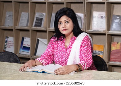 Indian woman reading book in college library with bookshelf behind her. - Shutterstock ID 2230682693