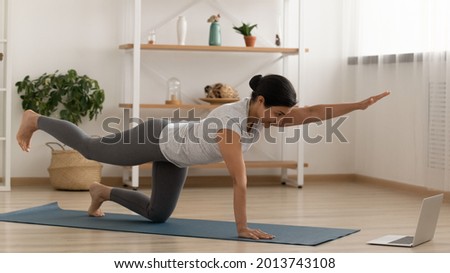 Indian woman in sportswear practicing yoga online, looking at laptop screen, doing Bird dog exercise on mat floor, sporty young female involved in internet lesson, watching webinar, training at home