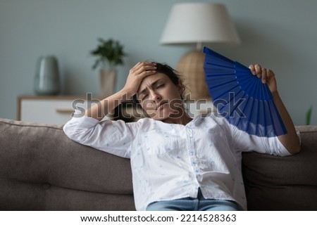 Indian woman suffers from heat inside, cools herself waving blue paper fan, leaned on sofa in living room without air-conditioner, touch forehead feels unwell. Hormonal imbalance, hot weather indoors [[stock_photo]] © 