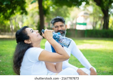 Indian Woman Take A Break While Sitting In The Park Drinking Water After Yoga Exercise Workout On A Hot Day, Asian Couple Relaxing In The Garden Together Outdoor. Selective Focus