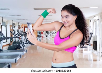 Indian Woman Taking A Selfie Picture With A Smartphone While Showing Her Arm Muscle. Shot In The Gym Center