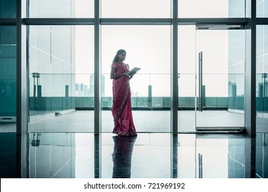Indian woman using a tablet on the corridor of a modern corporate building with tiled floor and transparent walls